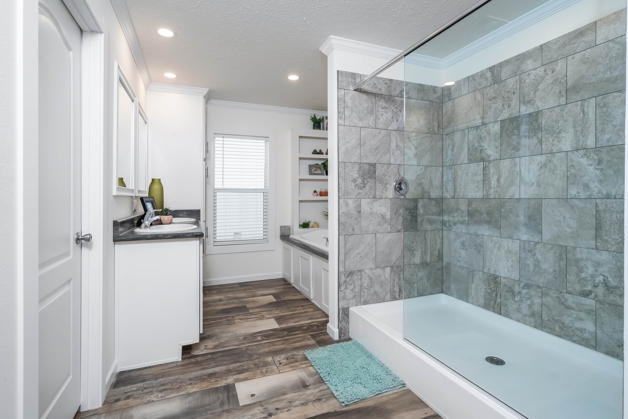 Spacious bathroom with wood floor, large gray-tiled shower, dual sinks with dark countertops, bathtub near the window, and shelves with decor.