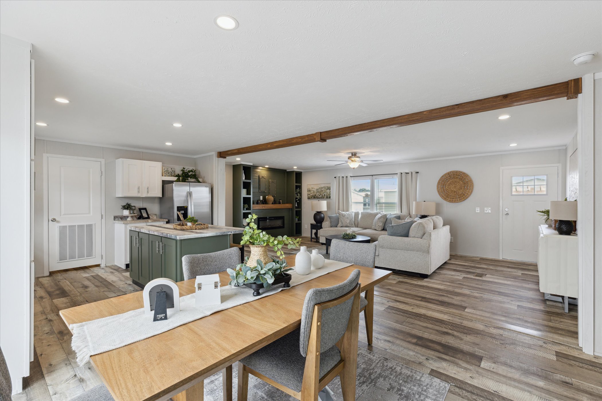 Open-concept living room and kitchen with modern decor. Wooden dining table, cozy sofas, green kitchen cabinets, and hardwood flooring create a welcoming atmosphere.