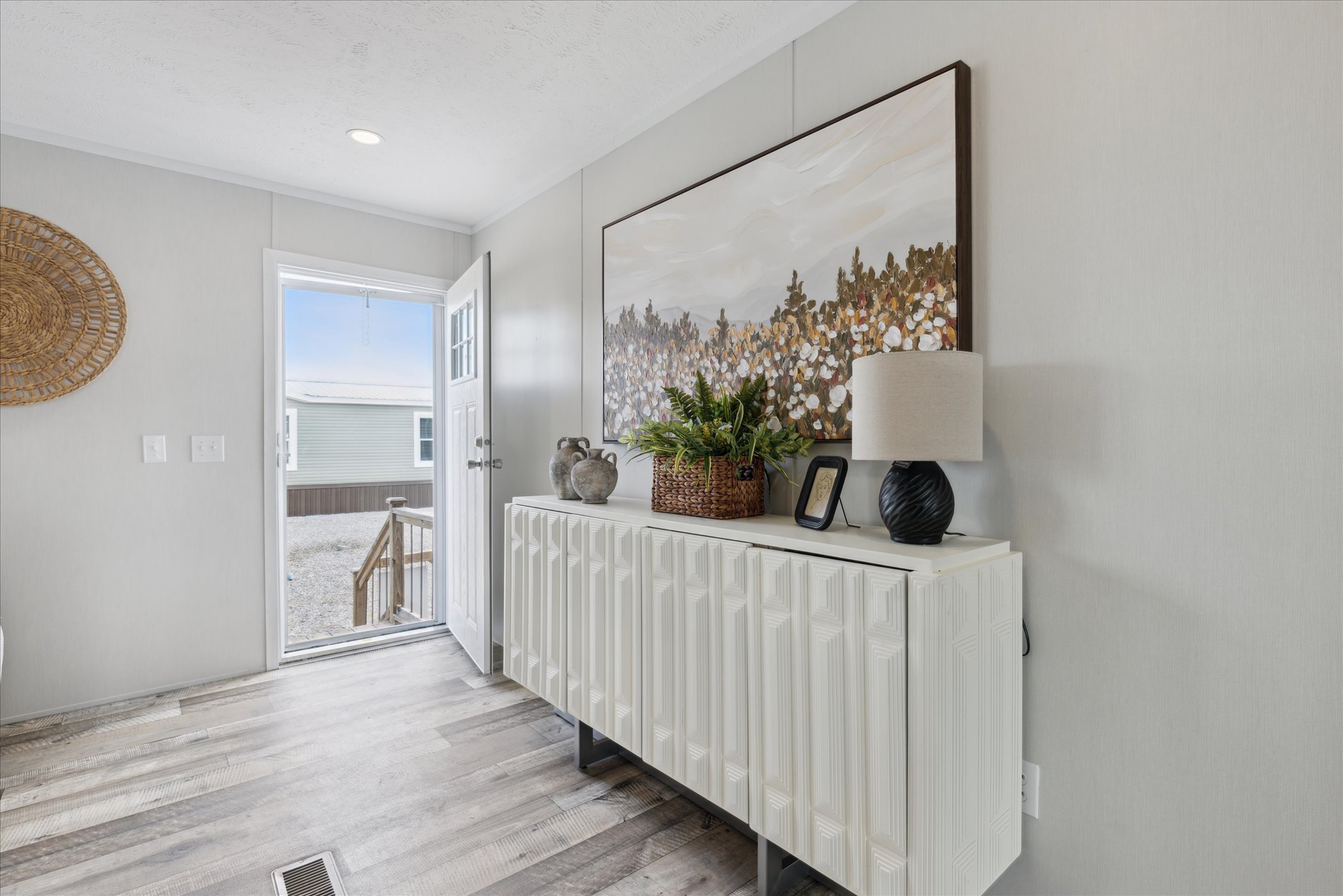 A bright foyer with a textured white cabinet, topped with a plant, lamp, and decor. Above is a landscape painting. Open door reveals outdoor stairs.