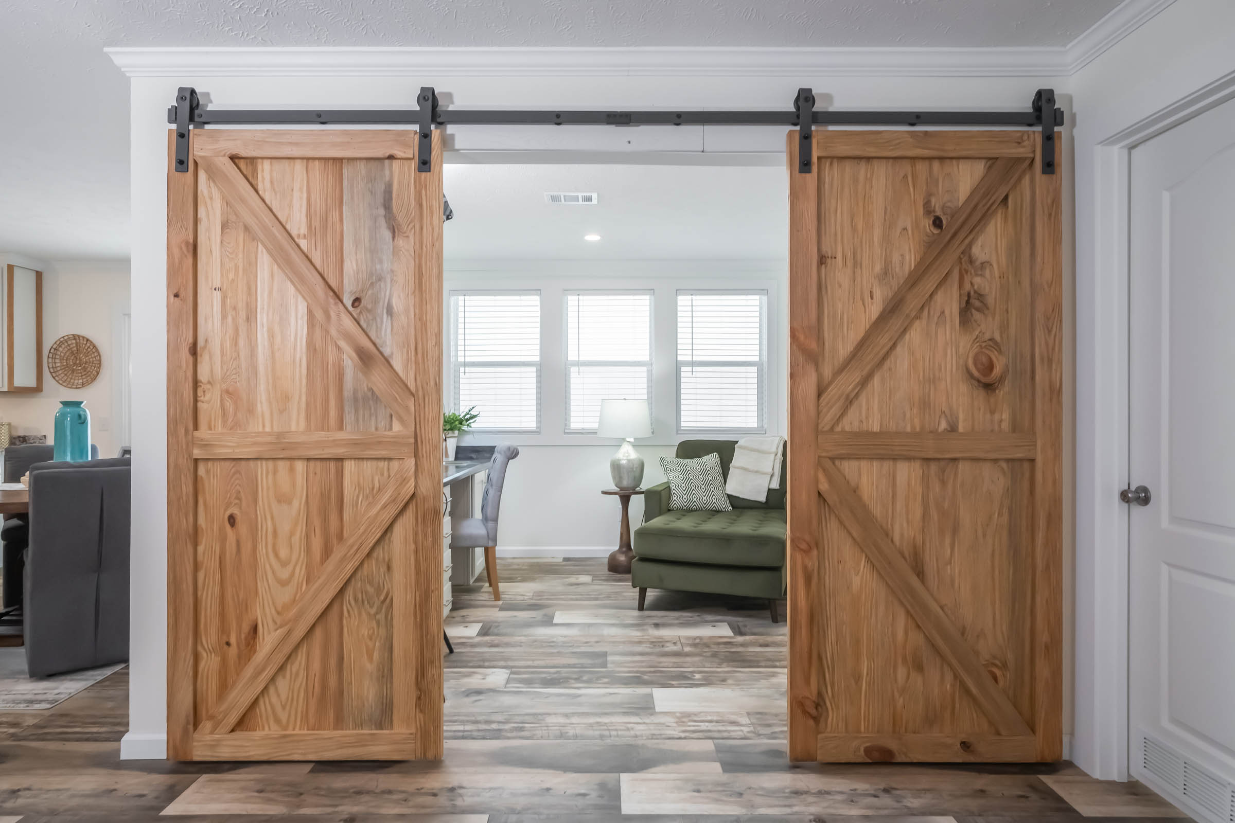 Natural wood sliding barn doors open to a cozy living room with an olive sofa, side table, and large windows, creating a warm, rustic ambiance.