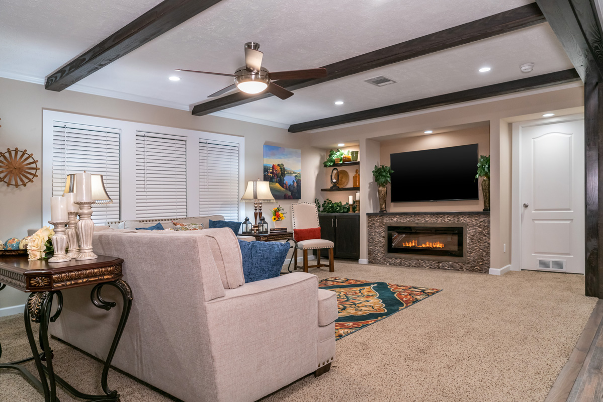 A cozy living room with beige walls and carpet, featuring a grey sectional sofa, patterned rug, and a modern fireplace beneath a large TV. There are wooden ceiling beams, a fan, and ambient lighting creating a warm and inviting atmosphere.
