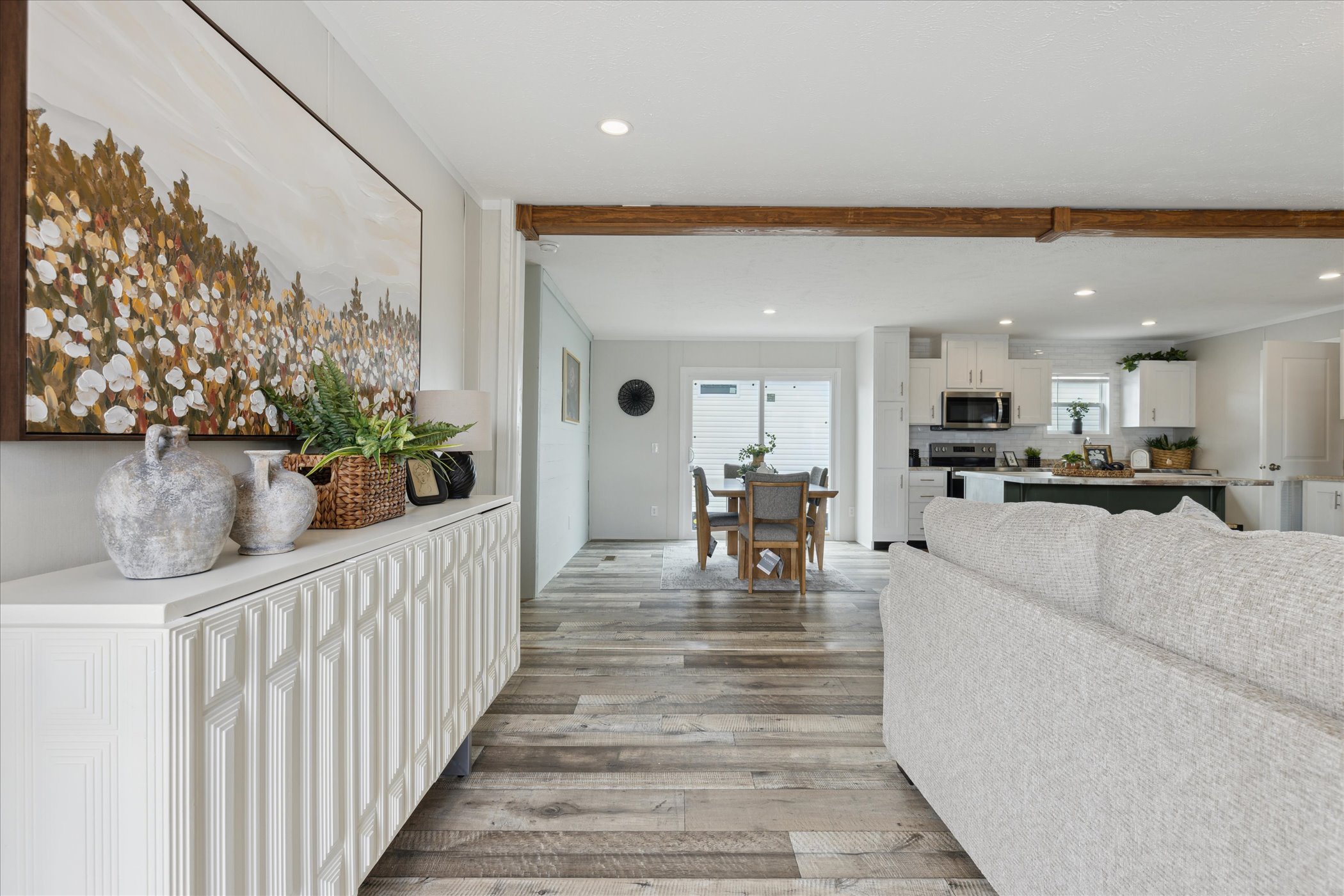 Spacious living room with light gray sofa, wood floor, large floral painting, and decorative vases. Dining area and modern kitchen in background. Warm, cozy atmosphere.