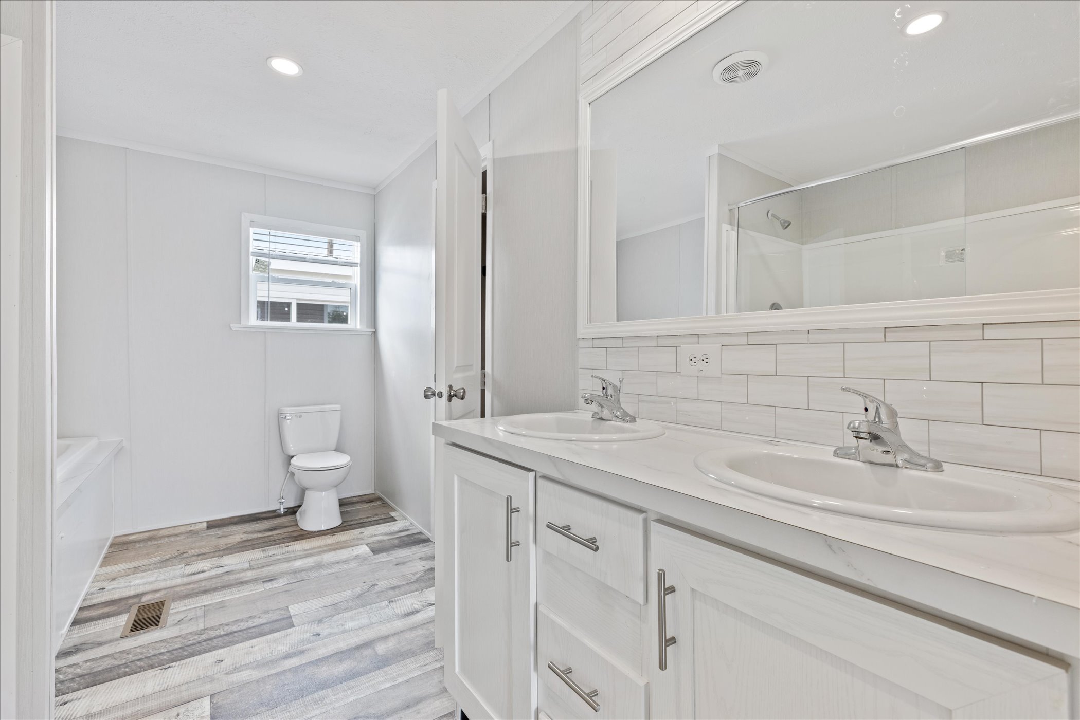 Bright, modern bathroom with dual sinks on a white vanity, large mirror, wood-style tile flooring, and a toilet near a small window. Clean and minimalist.