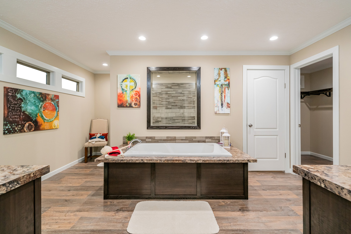 Modern bathroom with a central bathtub, beige walls, and wood flooring. Colorful abstract art decorates the walls, and recessed lighting sets a calm tone.