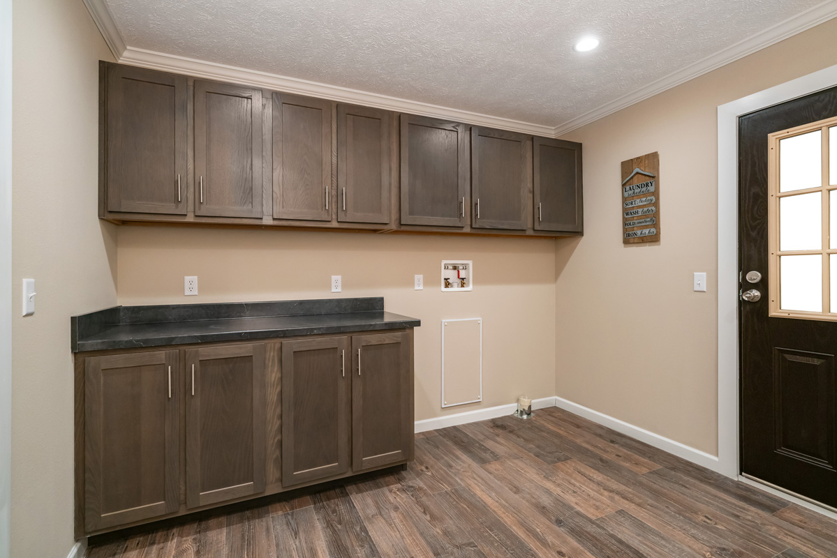 Laundry room with dark wood cabinets, beige walls, and wood flooring. A small sign hangs on the wall near a black door with a window.