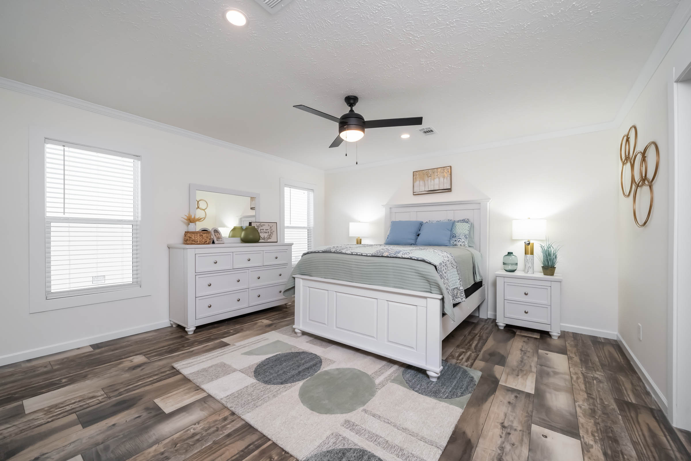 A bright, modern bedroom with white furniture, wood flooring, and a ceiling fan. The room features a bed with blue and green bedding, two nightstands, and a dresser with a mirror. Artwork and decor add warmth.