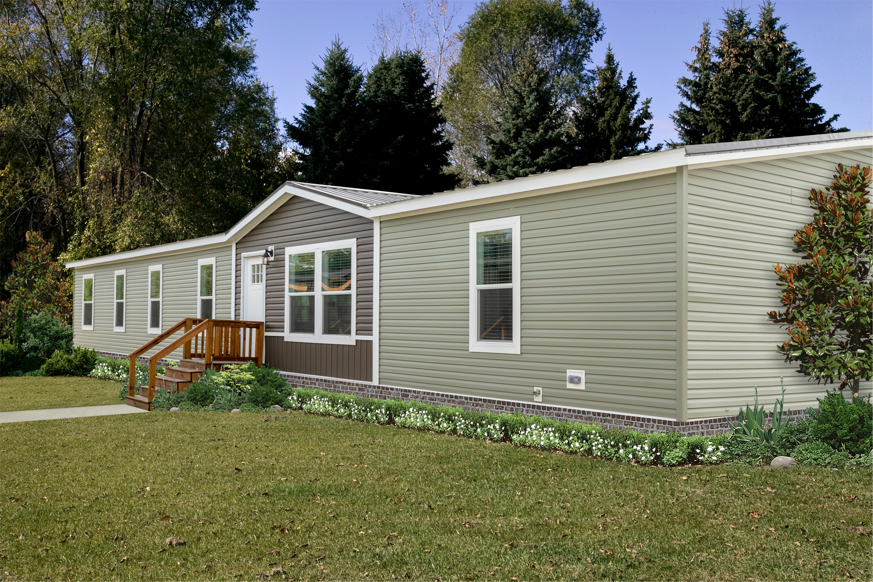 A beige, single-story manufactured home with white window frames sits on a well-kept lawn surrounded by trees. A small wooden porch leads to the entrance.