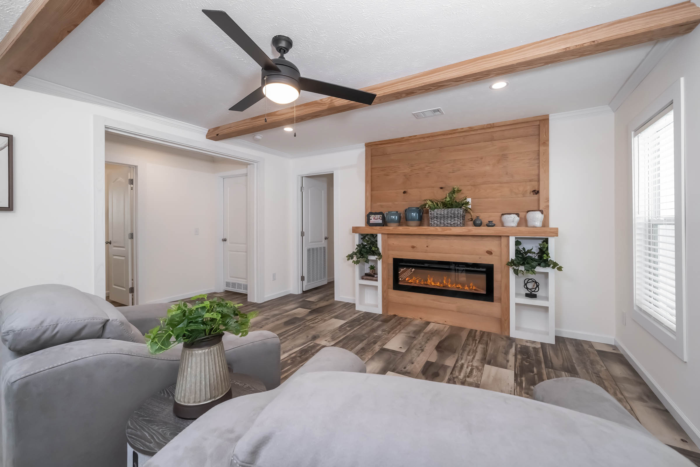 Modern living room with a ceiling fan, wooden beams, and an electric fireplace. Cozy gray furniture and plants create a warm, inviting ambiance.